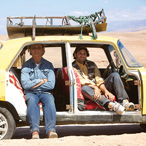 Two men sit in an old, yellow car with the doors open in a desert landscape. One sits in the drivers seat, the other in the back. The car has items tied on the roof and red patterned seat covers.