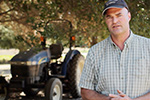 A man wearing a plaid shirt and baseball cap stands outdoors in front of a blue tractor, with trees providing shade in the background.
