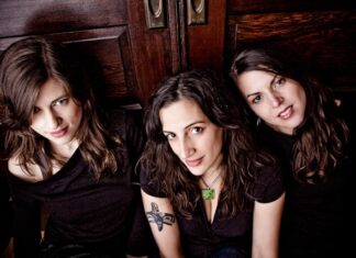 Three women with long brown hair sit closely together in front of a wooden door, all wearing dark tops. One has a visible tattoo on her upper arm. They face the camera and smile softly.