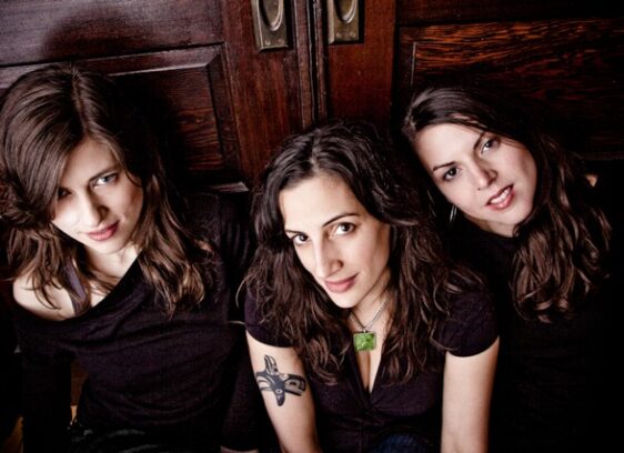 Three women with long brown hair sit closely together in front of a wooden door, all wearing dark tops. One has a visible tattoo on her upper arm. They face the camera and smile softly.