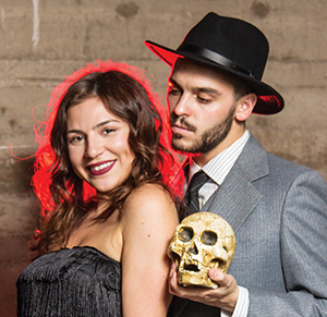 A woman in a black dress smiles at the camera while a man in a suit and hat stands beside her, holding a skull and looking at it. The background is a textured wall with red lighting.