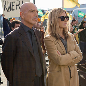A bald man in a plaid suit and a woman with blonde hair in a tan coat, both wearing sunglasses, stand side by side at an outdoor event with people holding signs and flags in the background.