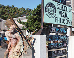 A sculpture of a partially abstract human figure stands next to a sign for the School of Visual Philosophy, with several workshop listings and trees in the background on a sunny day.