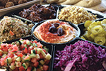 A platter with sections of various Mediterranean dishes, including hummus topped with olives, tabbouleh, chopped salad, pickles, shredded red cabbage, caramelized onions, and couscous.