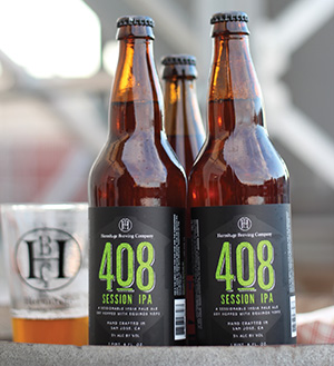 Four brown glass bottles of 408 Session IPA beer with black labels and green text are grouped together next to a clear plastic cup partially filled with beer. The background is softly blurred.