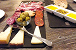 A charcuterie board with assorted cheeses, salami, cured meats, green olives, crackers, and a fork, along with a tray of dipping oils and sliced bread on a wooden table.