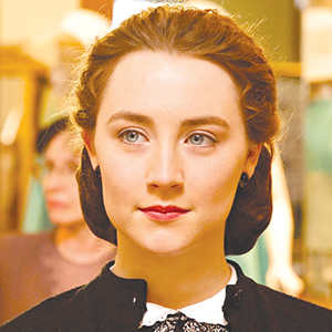 A young woman with fair skin, blue eyes, and auburn hair styled back, wearing a black top with a white collar, stands indoors with a soft, focused expression. There are blurred figures and warm lighting in the background.