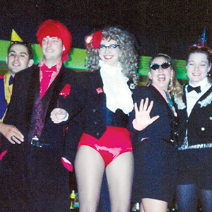 A group of five people dressed in colorful, playful costumes, including party hats, sunglasses, a bright red wig, and red shorts, smiling and posing together at a festive event.