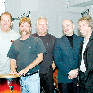 Five men stand closely together indoors, smiling. One man on the left rests his hands on a drum, and the others are dressed casually or in jackets, posing in a music studio setting.
