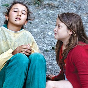 Two young girls sit on the ground outdoors. One girl, wearing a yellow top and green pants, has her eyes closed and a black mark near her mouth. The other girl, in a red sweater, looks at her attentively.