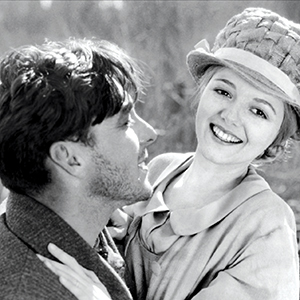 A black and white photo of a young man and woman outdoors. The man, in profile, looks at the woman, who is smiling brightly and wearing a textured hat and coat. They appear happy and close together.