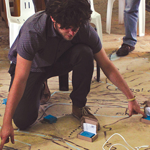 A person crouches on the ground, arranging small blue electronic devices connected by wires on a large map. White chairs and another person standing are visible in the background.