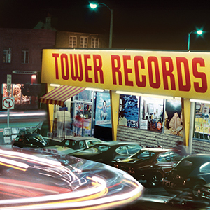 A brightly lit Tower Records store at night, with a large yellow and red sign above the entrance, several parked cars in front, and blurred streaks of car lights in the foreground.