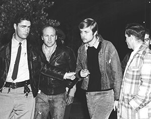 Four men stand together in a black and white photo, wearing jackets and jeans. One man appears to be in handcuffs, escorted by another man in a police uniform, while others look on. Trees are visible in the background.