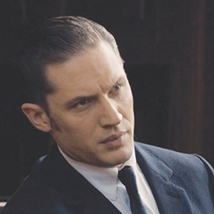 A man with slicked-back hair wearing a dark suit, white shirt, and tie looks to the side with a serious expression in a dimly lit setting.