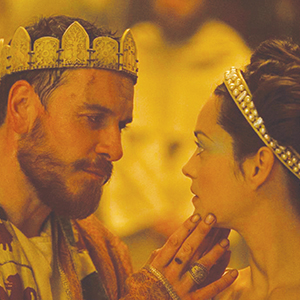 A man wearing a gold crown gently touches the chin of a woman with a jeweled headband. They gaze intensely at each other, dressed in ornate, medieval-style clothing, in a warmly lit setting.