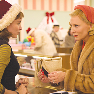 Two women in vintage clothing talk at a store counter during the holidays; one wears a red coat and headband, the other a Santa hat. Holiday decorations and shoppers are visible in the background.