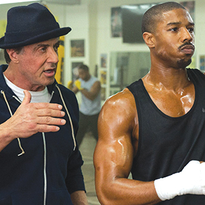 A man in a hat and hoodie gestures while talking to a muscular, sweaty man in a black tank top with his hands wrapped, standing in a gym with boxing equipment in the background.