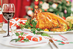 A festive table setting with a roasted turkey, a glass of red wine, wrapped gifts, and holiday decorations. A Christmas tree with lights is blurred in the background.