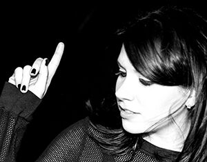 A woman with dark hair and a mesh top looks down and to the side, raising one hand with her index finger pointed upward against a black background. The image is in black and white.