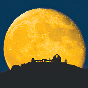 A large, bright full moon rises behind the dark silhouette of an observatory and trees on a hilltop, set against a deep blue night sky.