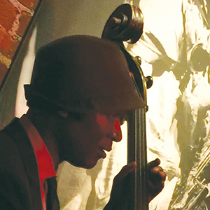 A musician wearing a hat plays an upright bass in a dimly lit room with a large black-and-white artwork visible in the background.