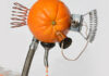 An orange with attached metal tubes and coils dispenses juice through a spout into small cups arranged in a row below it.