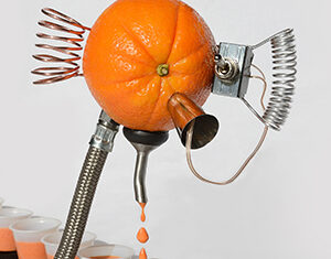 An orange with attached metal tubes and coils dispenses juice through a spout into small cups arranged in a row below it.