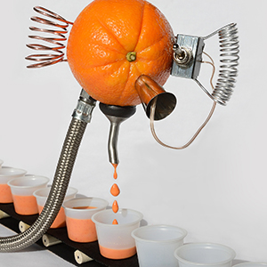 An orange with attached metal tubes and coils dispenses juice through a spout into small cups arranged in a row below it.