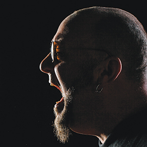A man with a shaved head, beard, and sunglasses is shown in profile, yelling or singing with his mouth wide open against a dark background.