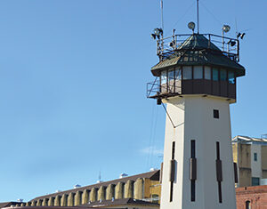 A tall, white lighthouse with a hexagonal observation deck stands in front of a large, tan building complex under a clear blue sky.