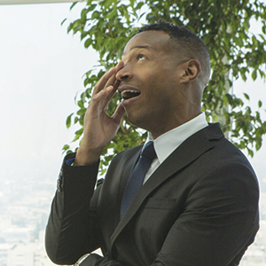 A man in a suit stands indoors, smiling and talking on his phone, with one hand on his face. There is a large green plant and a bright window in the background.