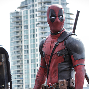 A person dressed in a red and black superhero costume with a mask and sword stands outdoors in front of a modern high-rise building.