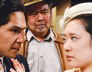 Three people stand close together. A man and woman face each other intensely while a third older man stands behind them, watching with a serious expression. The woman wears a white hat and outfit; the men wear darker clothing.