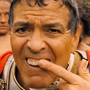 A man dressed in a Roman-style costume points to his teeth and grimaces, with other people blurred in the background.