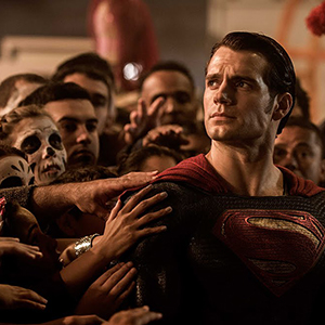Superman stands surrounded by a crowd of people reaching out to touch him; some have painted faces, and he looks ahead with a serious expression, wearing his iconic suit with a red and yellow S emblem.