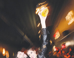 A woman smiling and holding a drink up high, with her arm around a man, in a dimly lit, crowded indoor setting. The image has a blurred effect, suggesting motion and a lively atmosphere.