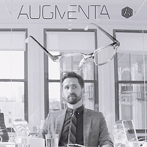 A man in a suit sits at a desk in a modern office. Above him, a large pair of eyeglasses and the word AUGMENTA are displayed, suggesting an augmented reality concept.