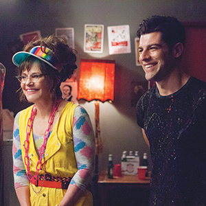 A woman in a colorful outfit and visor stands smiling next to a man in a black shirt. They are indoors, with a red lamp and party decorations in the background. Both appear happy and relaxed.
