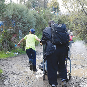 Two people walk through a muddy path; one pulls a cart loaded with bags while the other guides ahead, surrounded by trees and bushes.