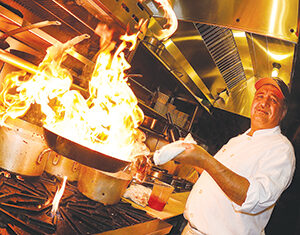 A chef in a white uniform and red cap cooks over a gas stove, creating tall flames in a pan inside a commercial kitchen. He smiles while holding the pan with one hand and a towel in the other.