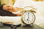 A blurred woman sleeping in bed with a gold analog alarm clock and a smartphone with earphones on a bedside table in the foreground.