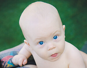 A baby with bright blue eyes rests on an adults chest, who has a colorful tattoo of a bird and flowers on their upper chest. The scene is outdoors with green grass in the background.