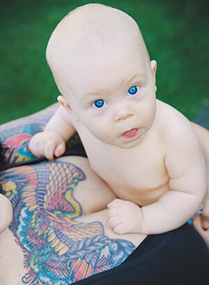 A baby with bright blue eyes rests on an adults chest, who has a colorful tattoo of a bird and flowers on their upper chest. The scene is outdoors with green grass in the background.