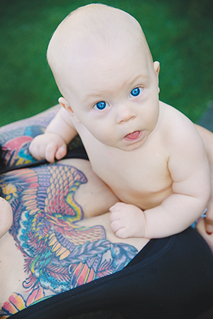 A baby with bright blue eyes rests on an adults chest, who has a colorful tattoo of a bird and flowers on their upper chest. The scene is outdoors with green grass in the background.