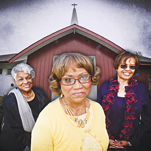 Three older women stand smiling in front of a church building with a cross-topped steeple. The woman in the foreground wears glasses and a yellow sweater; the two women behind her are dressed in dark clothing and also smiling.