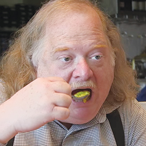 A man with long light brown hair and a mustache is wearing a checkered shirt and eating food with a fork. He is looking slightly to the side while taking a bite.