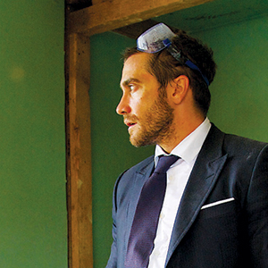 A man in a suit and tie stands indoors, looking to the side, with safety goggles resting on his head. The background is green with exposed wooden beams.