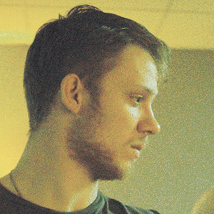 A young man with short brown hair and a beard is shown in profile, looking to the right. The background is softly lit and out of focus, creating a warm, subdued atmosphere.