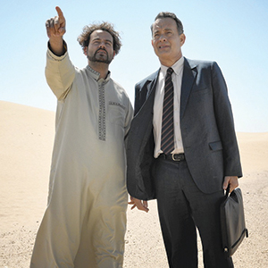 Two men stand in a desert under a clear blue sky; one wears a traditional robe and points ahead, while the other, dressed in a business suit and holding a briefcase, looks in the same direction.
